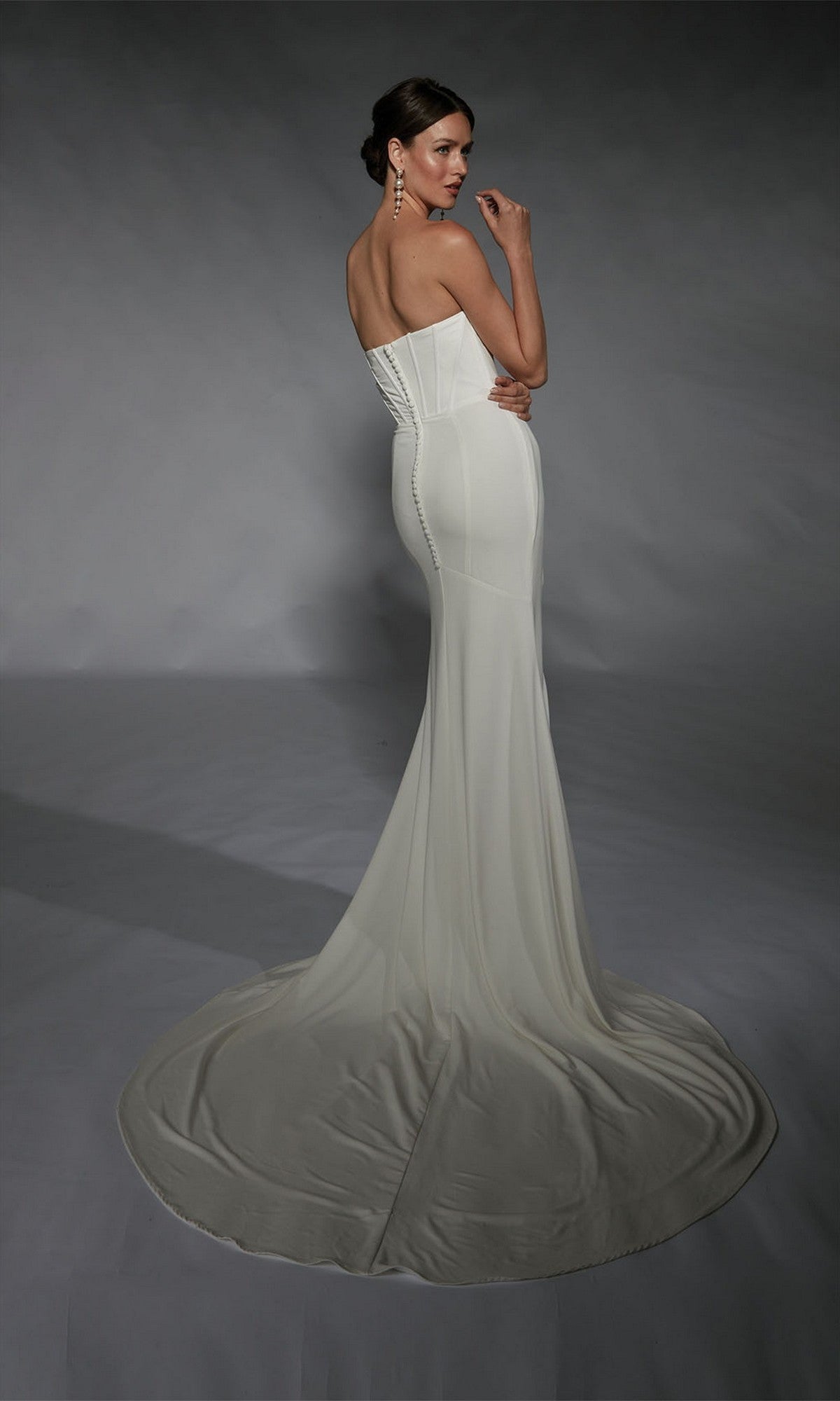 A woman wears the Long White Dress: Alyce 7102, featuring a minimalist design with a strapless corset bodice and long train, as she stands against a gray backdrop, facing away and looking over her shoulder.